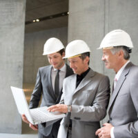 407cd22b6b6744cab7303a194c67af6e Businessmen in hard hats looking at colleagues laptop in office building